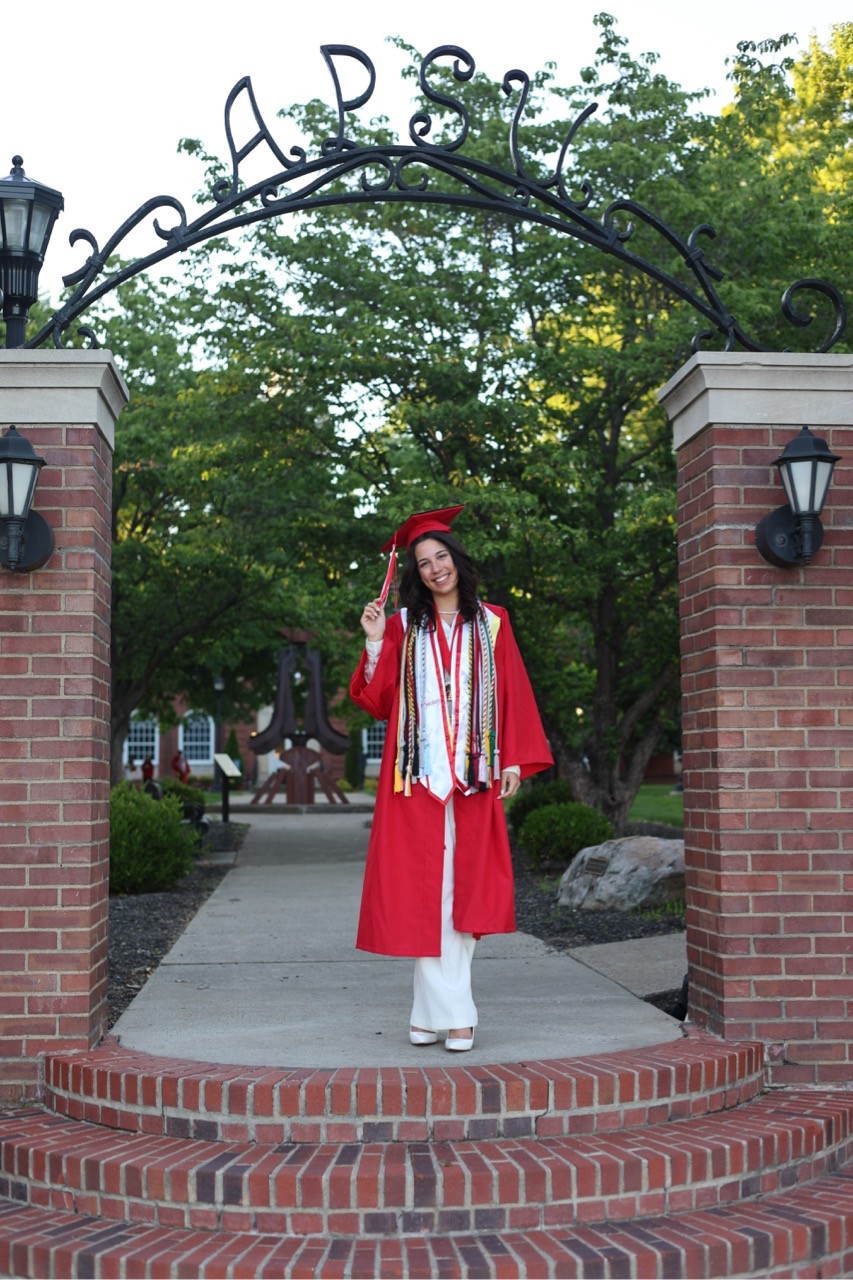 A smiling graduate in a red cap and gown stands under the ornate black metal "APSU" archway. She is wearing multiple colorful honor cords over a white jumpsuit and holding the tassel of her cap. The background shows a tree-lined campus path and brick pillars with lanterns.