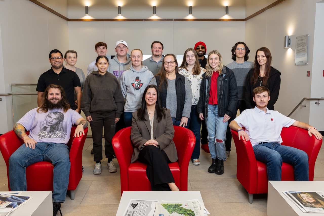 A group photo of approximately fifteen students and staff members in a bright, modern lobby. Three individuals are seated in vibrant red armchairs in the foreground; Daniela is in the center chair wearing a brown blazer. The rest of the group stands behind them in casual and business-casual attire.