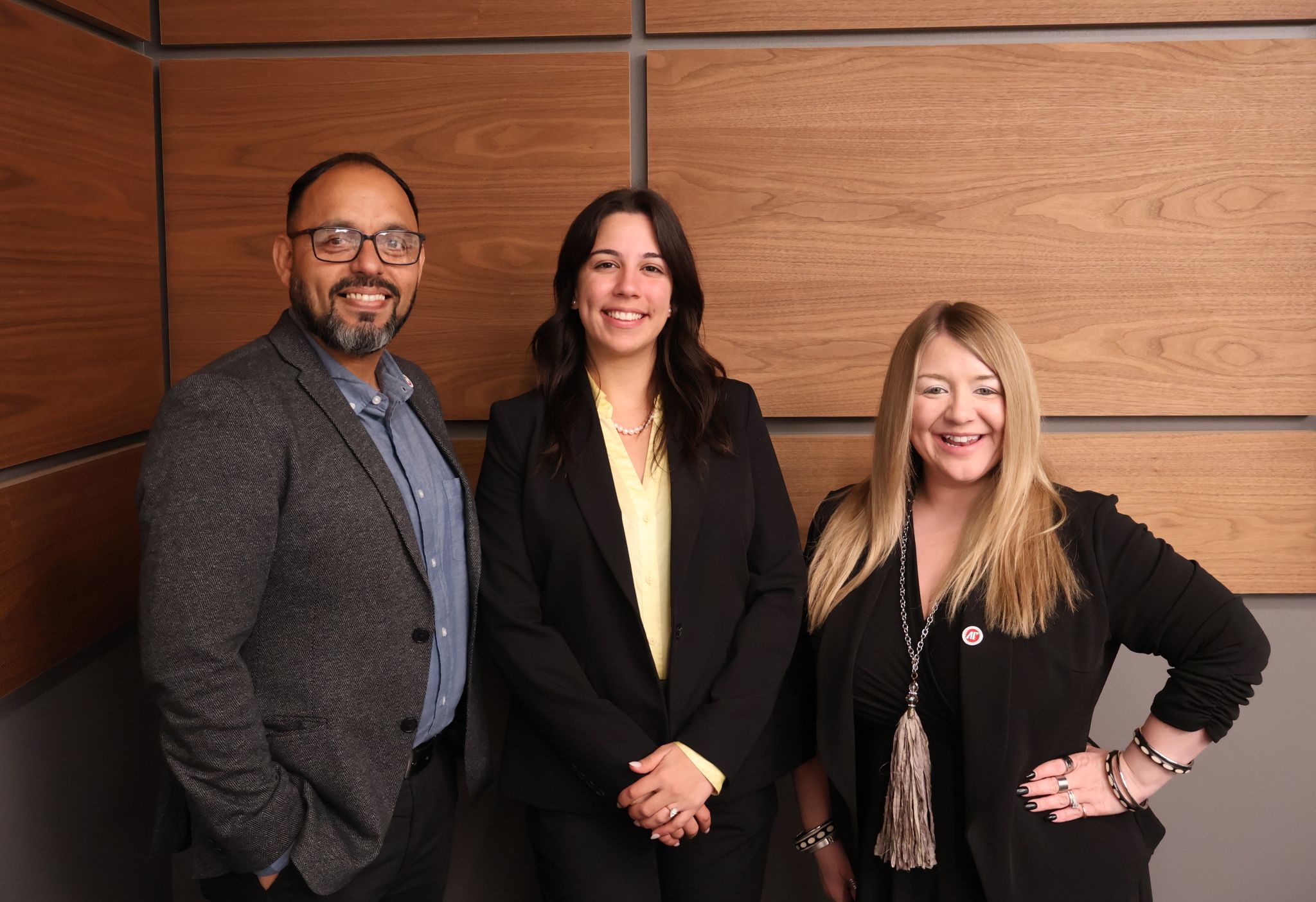 A professional portrait of three individuals posing in front of a wood-paneled wall. A man in a grey blazer stands on the left, Daniela is in the center wearing a black blazer and yellow blouse, and a woman in a black top with a long tassel necklace stands on the right.