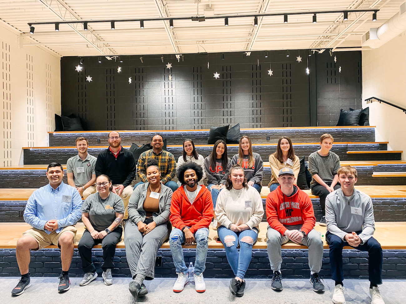 A group of about fifteen people sits on tiered wooden seating against a dark brick wall. The room is decorated with white paper snowflakes hanging from the ceiling. The group consists of students and faculty in a mix of casual clothing, including several wearing Austin Peay branded apparel.