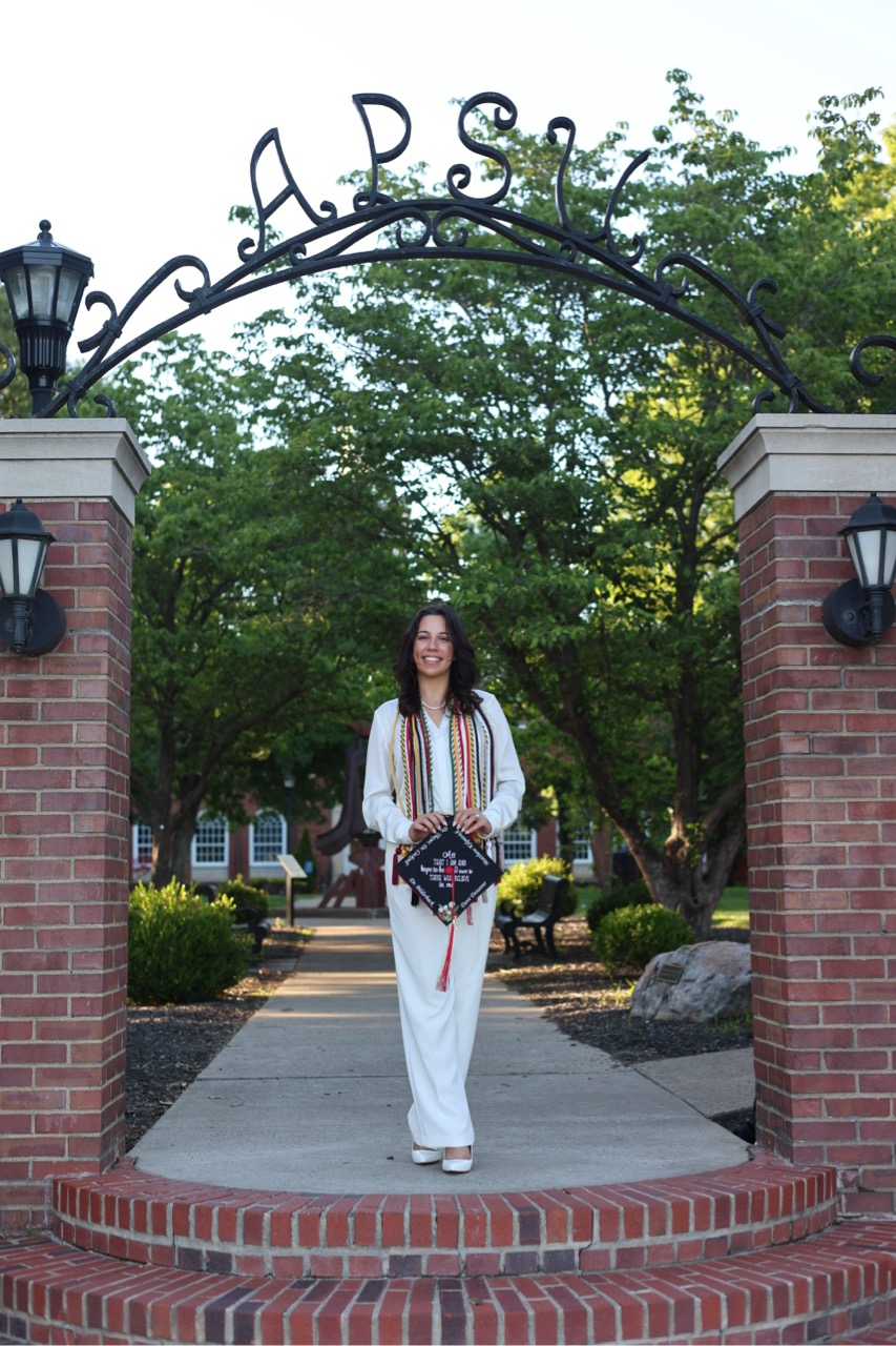 A graduate stands under the "APSU" iron archway wearing a white jumpsuit and several honor cords. She is holding her graduation cap, which is decorated with flowers and a personalized message. The setting is the same brick-lined campus entrance seen in the first photo.