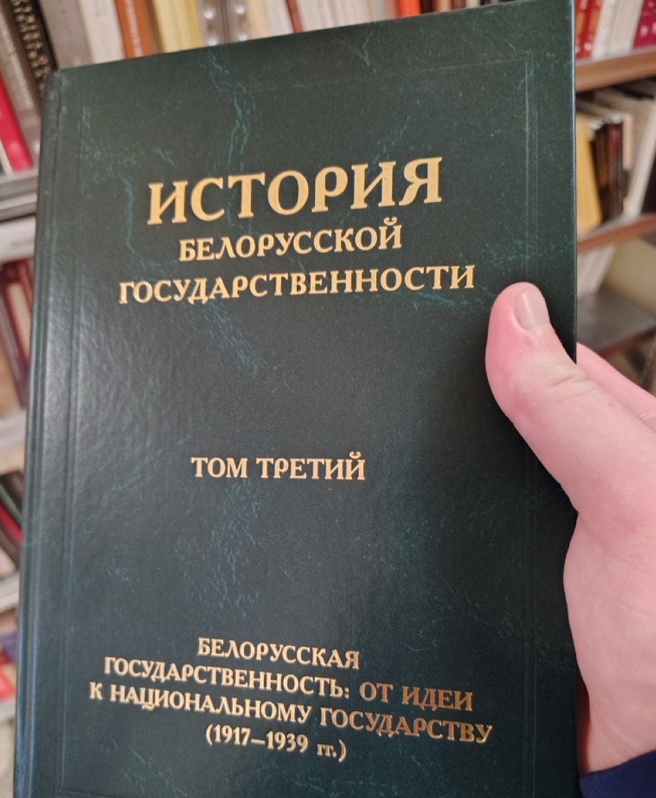 A hand holds a dark green hardcover book titled "History of Belarusian Statehood, Volume Three: Belarusian Statehood: From Idea to National State (1917–1939)" in Russian, with bookshelves visible in the background.