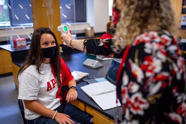 A professor, wearing a mask, takes a student's temperature in class.