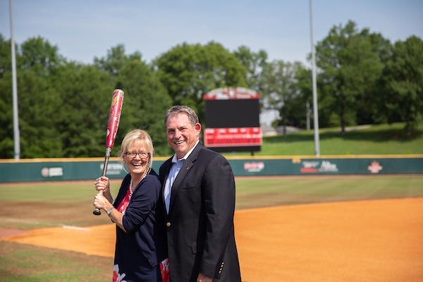 Doug and Linda Downey stand on a baseball field, while Linda holds a baseball bat.