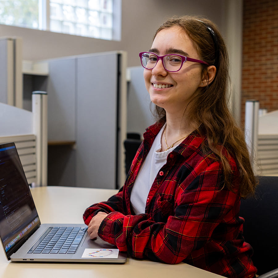 Female student in a red plaid shirt using a laptop at a cubicle desk and smiling at the camera.