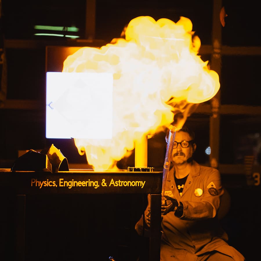 A professor in a lab coat kneels next to a table holding a clear tube with flames erupting from it during a demonstration, being lit up by the flames in a darkened room.