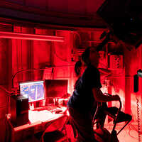 A student looks through a telescope in a dark, red-lit observatory with computers and equipment behind her.