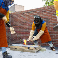 A student pouring molten metal into a mould while two other students stand to the sides holding extinguishers.