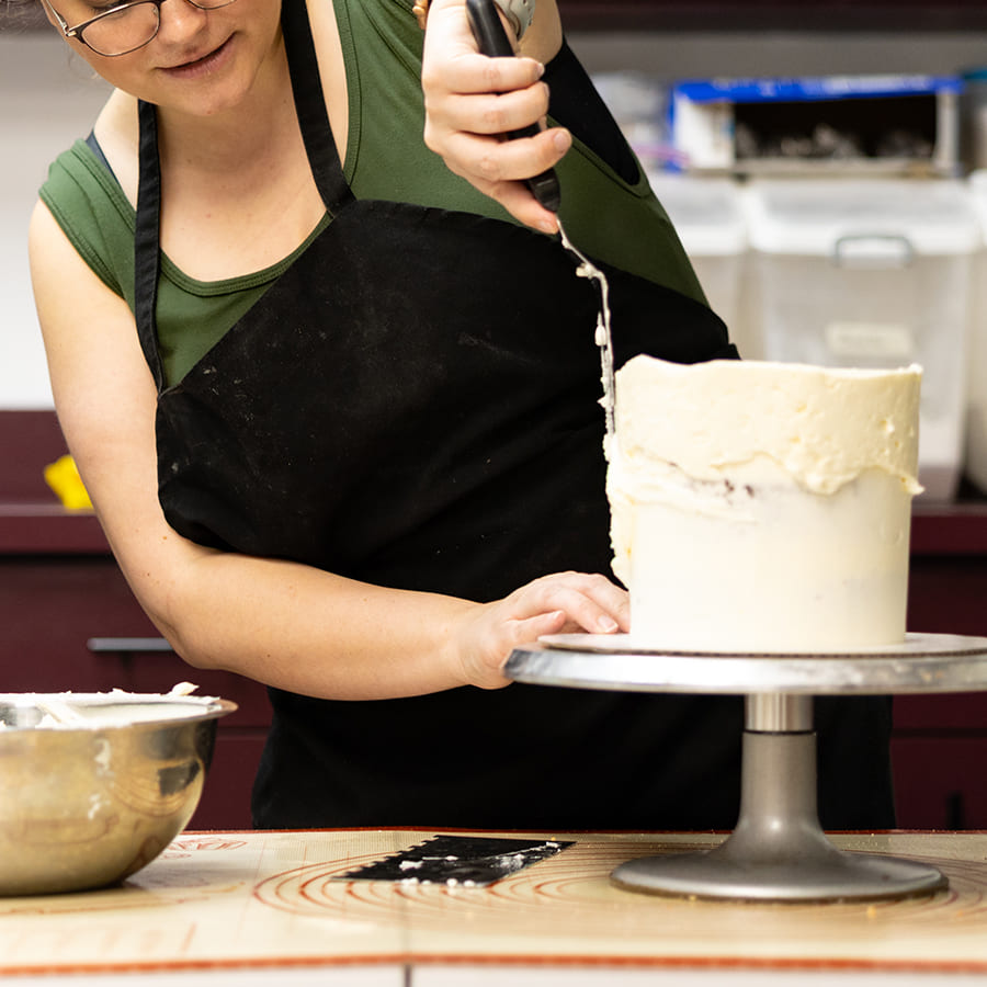 A baker frosts a white cake on a turntable, with a mixing bowl of icing beside them in a kitchen.
