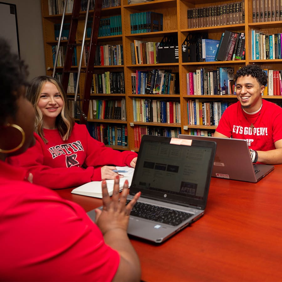 A group of four grad students sitting at a table in a library, engaged in discussion with open laptops and books.