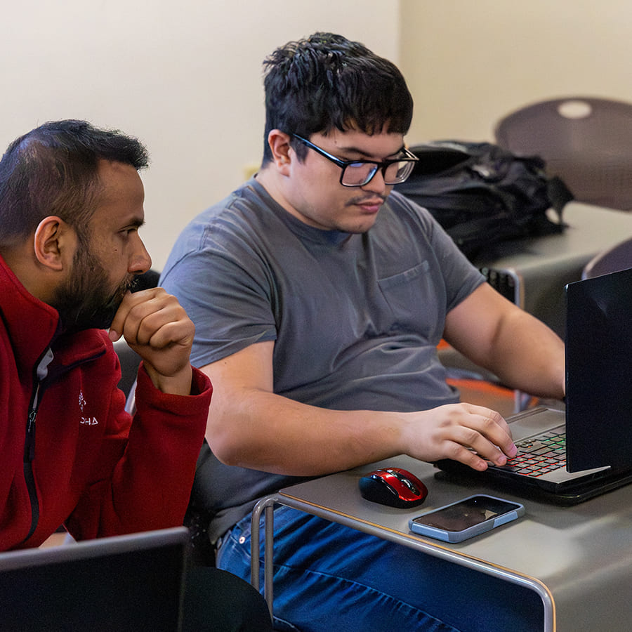 Two men working at a desk on a laptop in a classroom setting. One holds his chin thoughtfully, appearing focused and attentive, while the other types on the laptop.