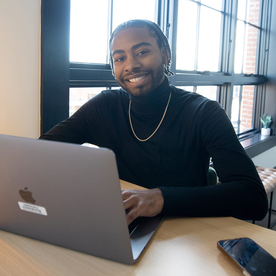 Smiling man wearing a black turtleneck and gold chain necklace, working on a laptop in a bright office.