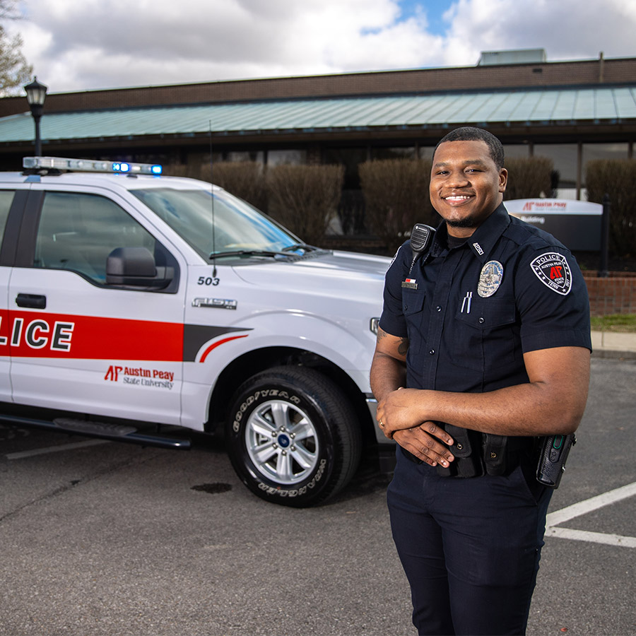 A police officer stands in front of a police truck with red and white markings.