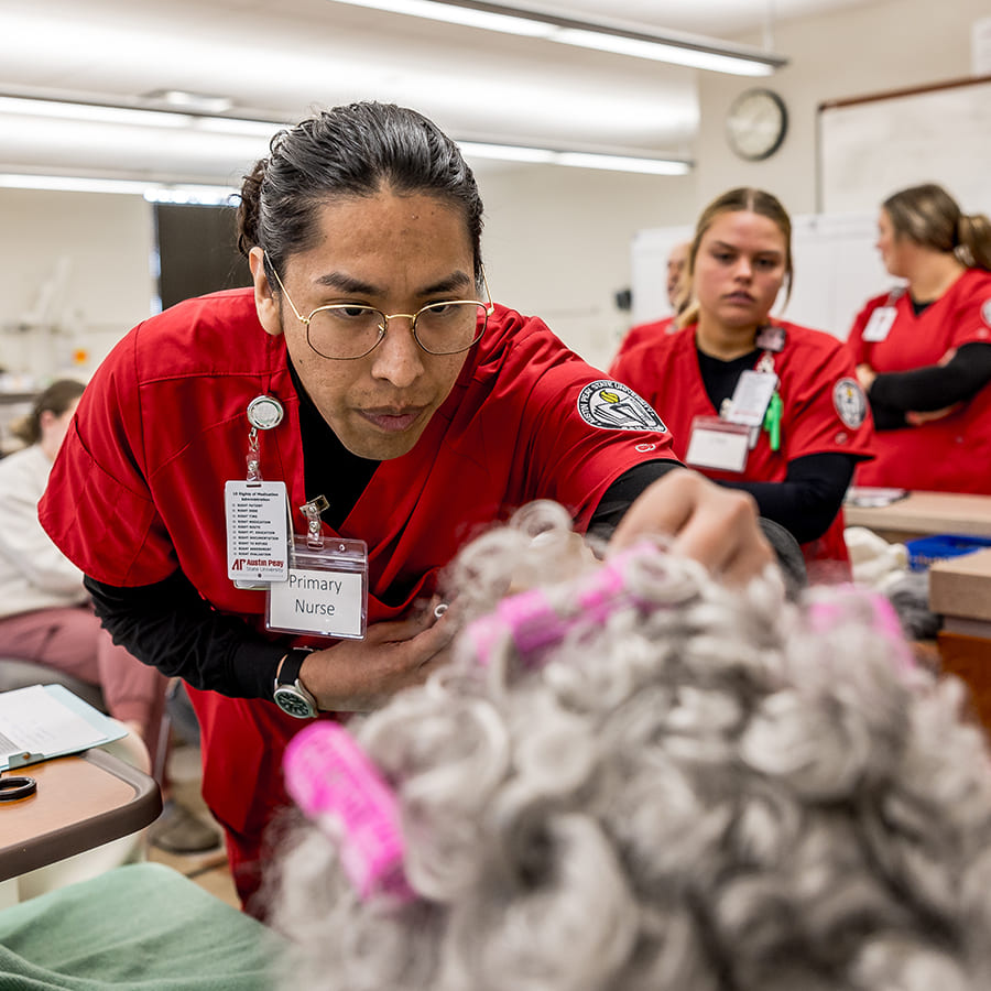 A nursing student examines an elderly mannequin during a training session in a simulated hospital, with other students in red scrubs observing.