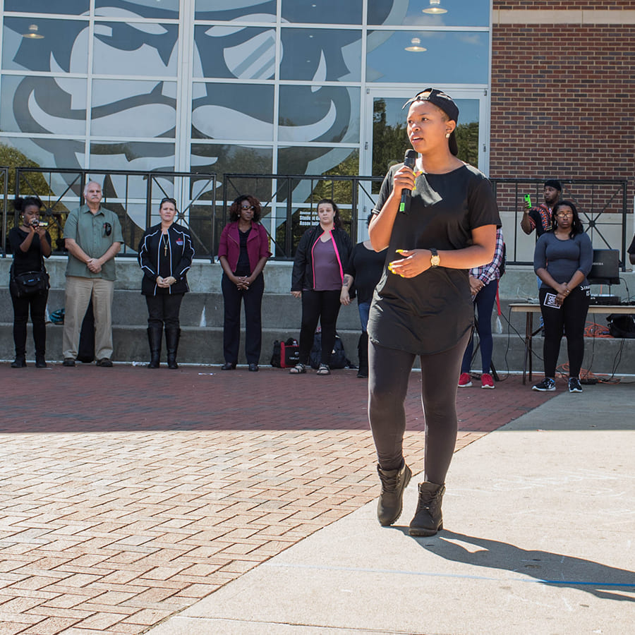 A group gathering outside of a large, diverse crowd gathered in a circle around a woman who is speaking confidently into a microphone.