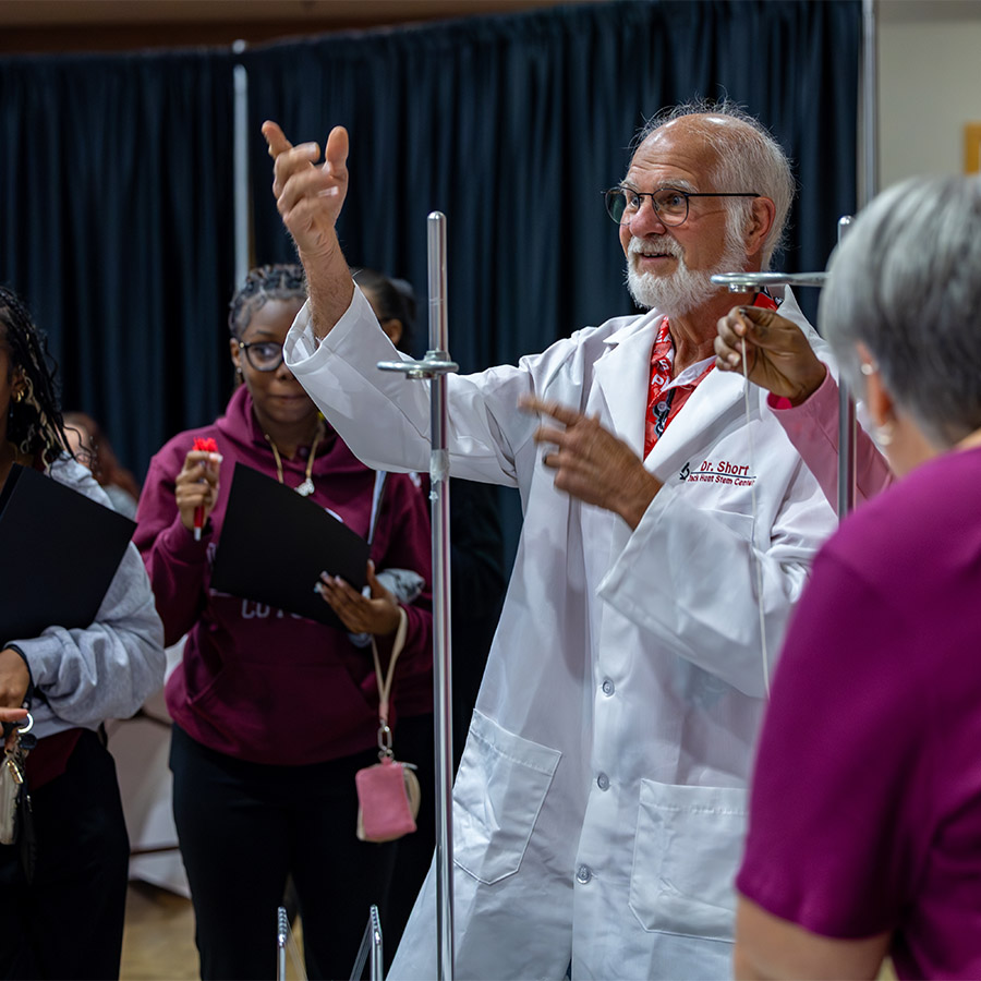 A science teacher in a white lab coat conducts a demonstration with a group of people watching attentively.