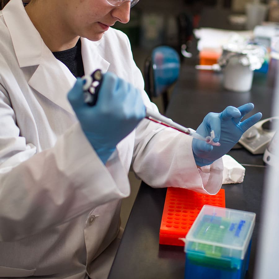 A student in a lab coat and blue gloves using a pipette in a laboratory setting.
