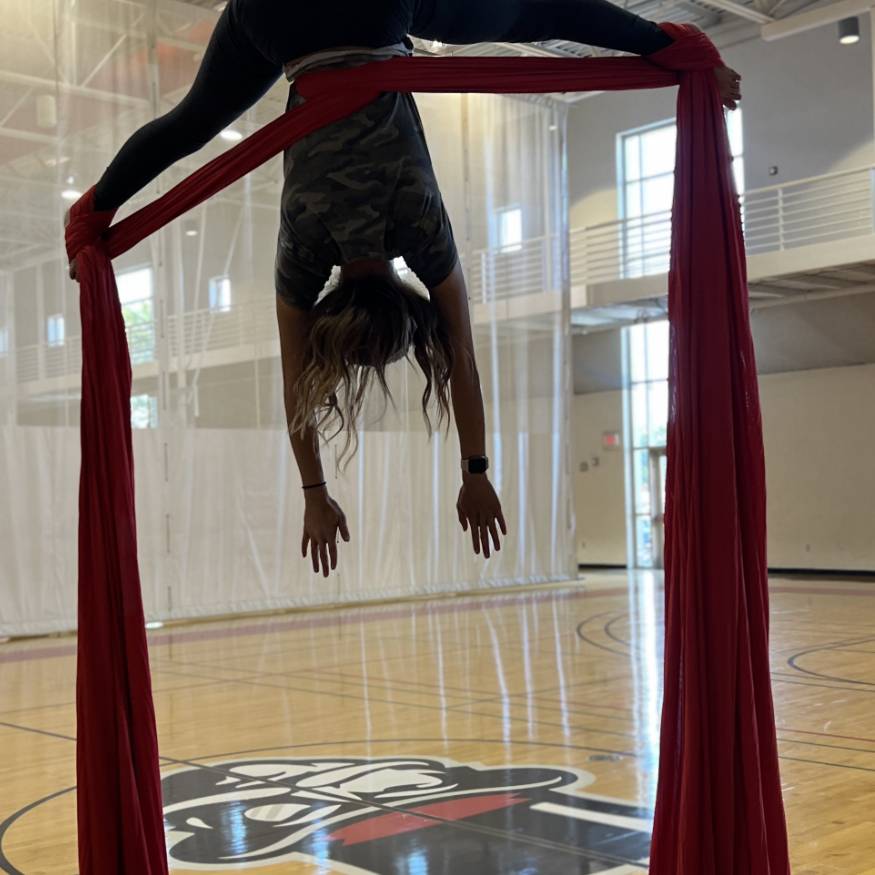 Aerial Silks student on red silk in crossback straddle, with gov head in background on gym court floor