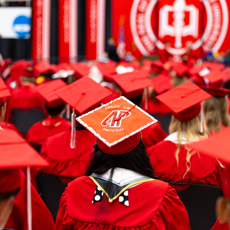 Rows of graduates in red caps and gowns seated at a commencement ceremony, with one mortarboard reading Trust the Process.