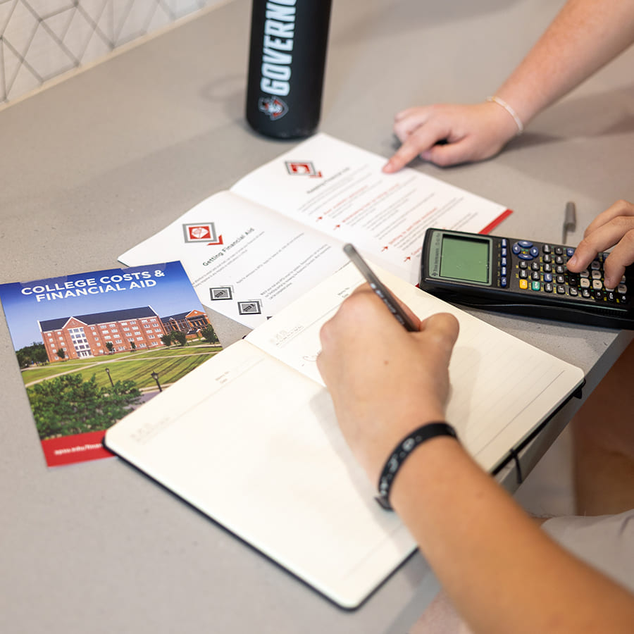 People reviewing financial-aid materials, writing in a notebook, and using a calculator at a table.