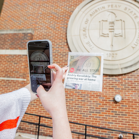 someone taking a picture of a newspaper in front of the Academic Seal on the side of the Morgan University Center