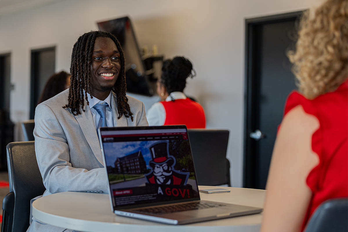 Smiling student in a light gray suit at a meeting table speaking with a staff member.
