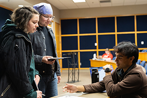 Two students speaking to a staff member who is seated at a desk with papers spread out in front of them.