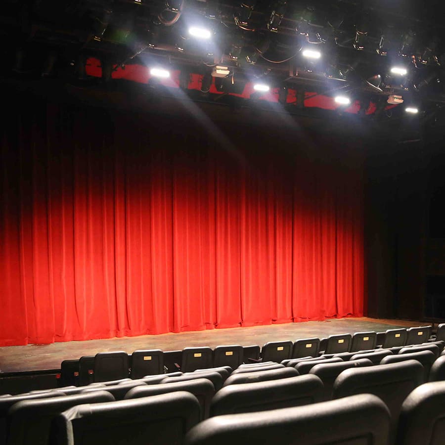 The empty Gotcher Theatre with the stage lights shining on the closed red curtain and empty seats in the foreground.