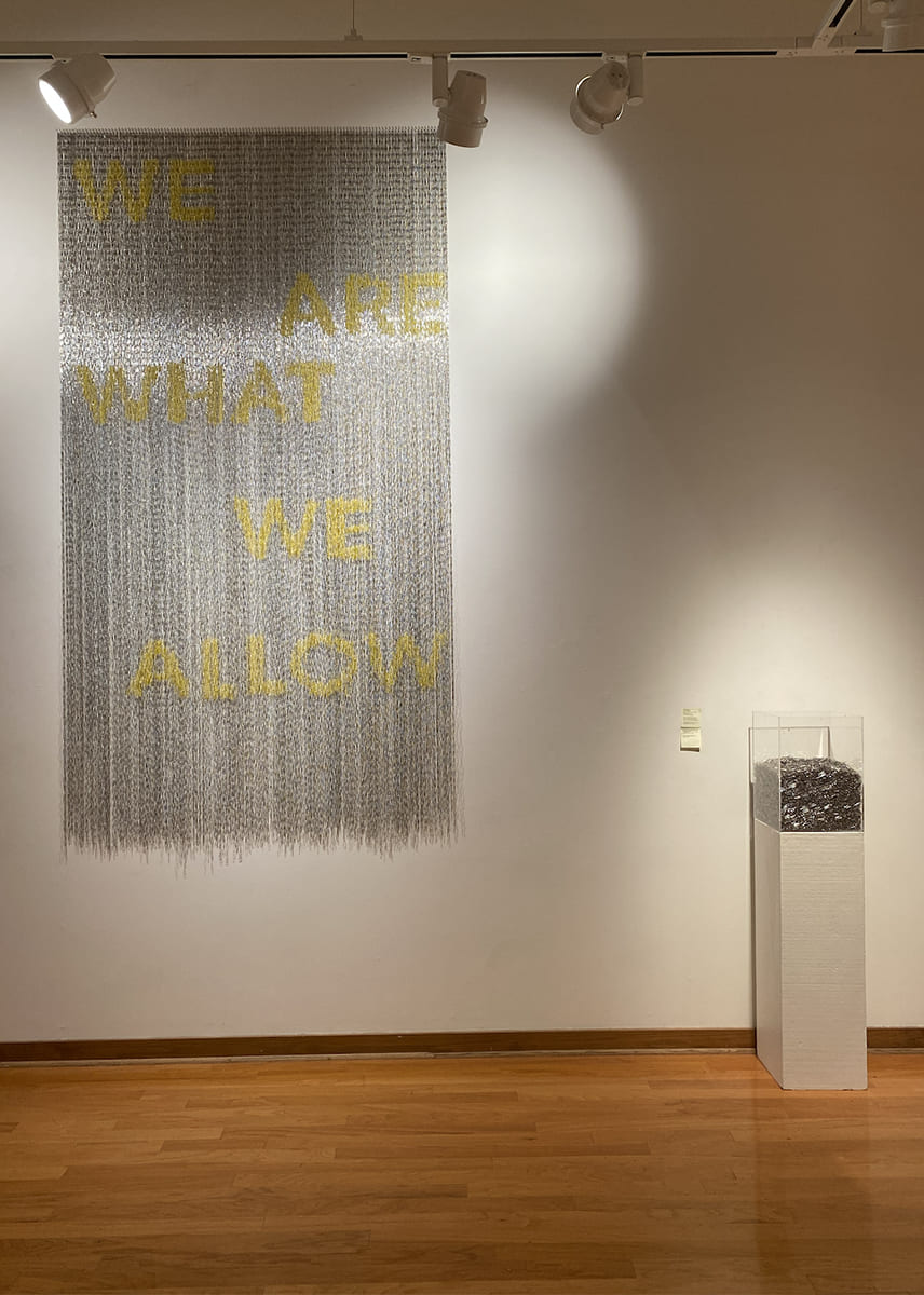 Suspended silver-gray paper-clip artwork with yellow text reading “WE ARE WHAT WE ALLOW,” displayed beside a pedestal case of paper clips.