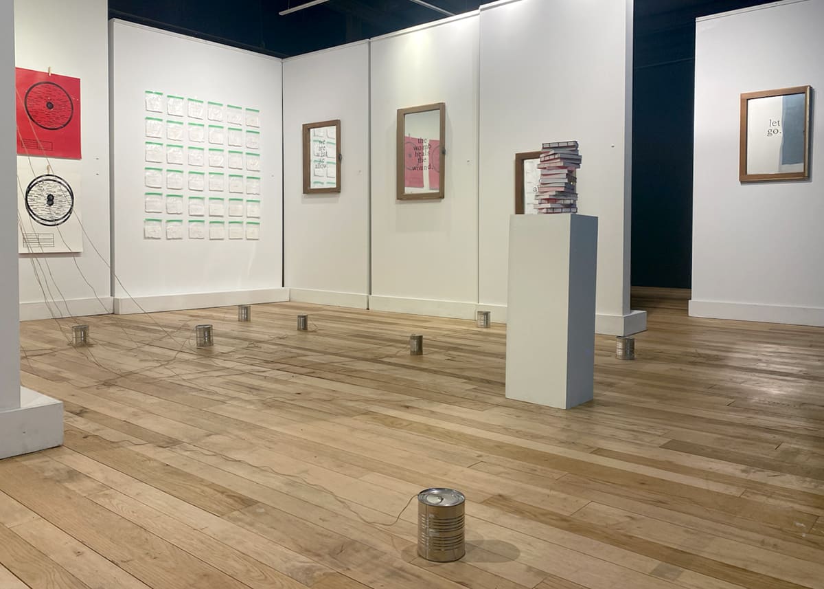 Installation view of Danielle Myers’s exhibition featuring framed text-based artworks, circular drawings, a stack of books, and metal cans connected by strings across a wood floor.