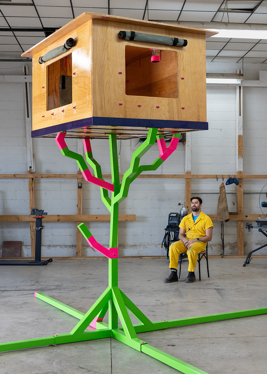 Artist Luke Warren sits in his studio beside a large wooden box sculpture elevated on neon green and pink branching supports.