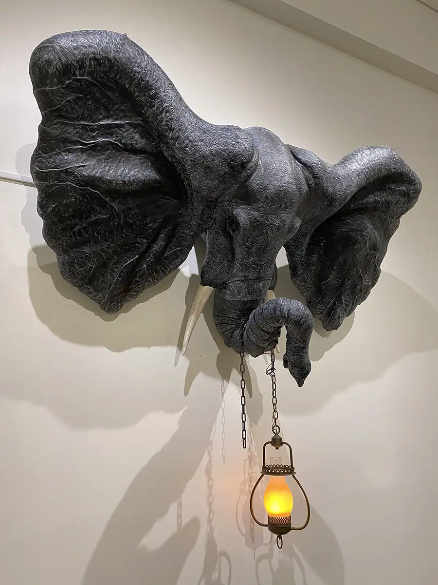 Stacey Holloway's artwork that features a detailed sculpture mounted on a wall, depicting the head of an elephant with large, textured ears and a prominent trunk. The sculpture is designed in a dark, matte shade, giving it a lifelike appearance. The elephant's trunk is extended downwards, holding a vintage-style lantern adorned with metal chains. The lantern emits a warm yellow glow that casts soft shadows on the wall, creating an atmospheric effect.