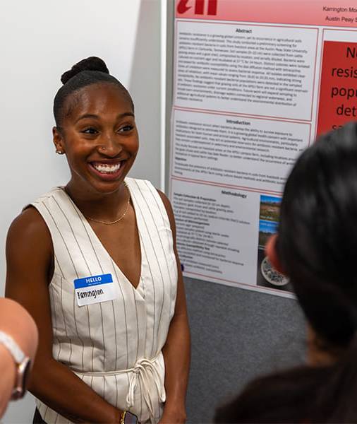 A student smiling broadly while presenting her research board to a small group.